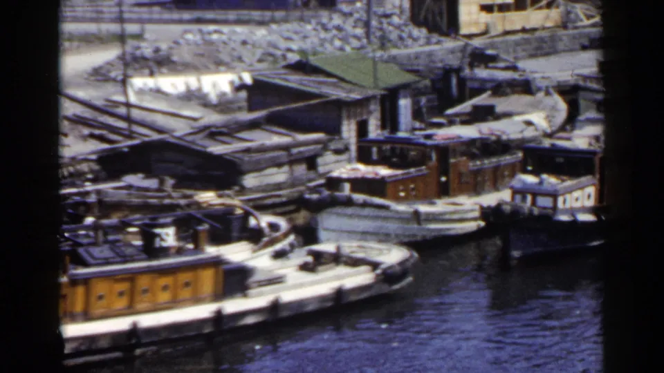 1953 Japan 8mm frame showing cabin boats clustered along the river with corrugated-roof structures and uneven shoreline behind.