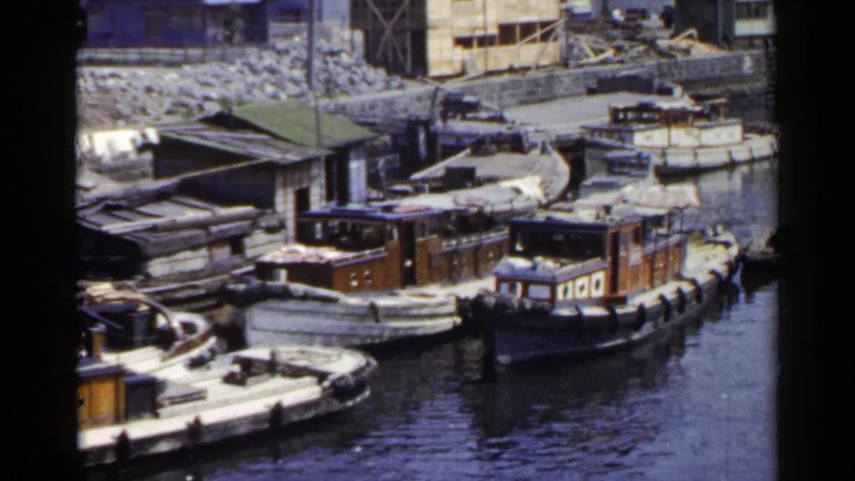 1953 Japan home-movie view of multiple wooden boats at a riverside landing with sheds, debris, and uneven bank conditions visible.