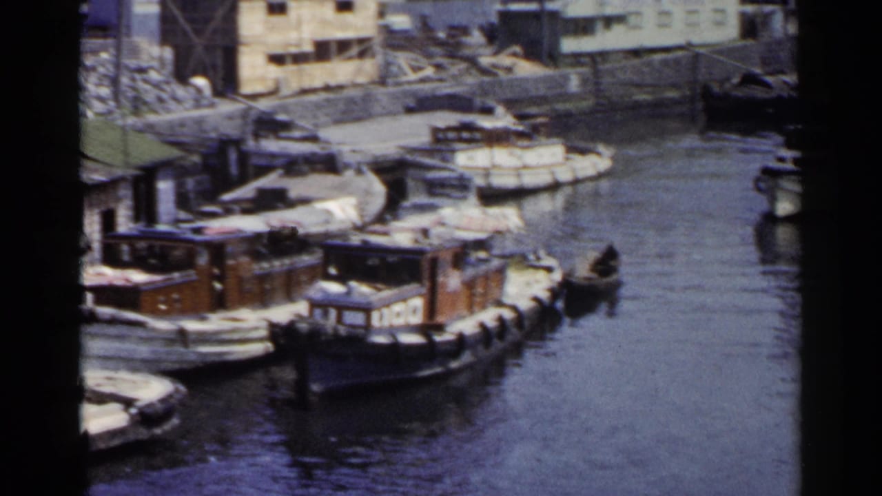 A 1953 Japan harbor view shows several small boats docked beside practical waterfront buildings, with a small dark craft on the water.