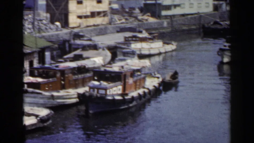 A 1953 Japan harbor view shows several small boats docked beside practical waterfront buildings, with a small dark craft on the water.