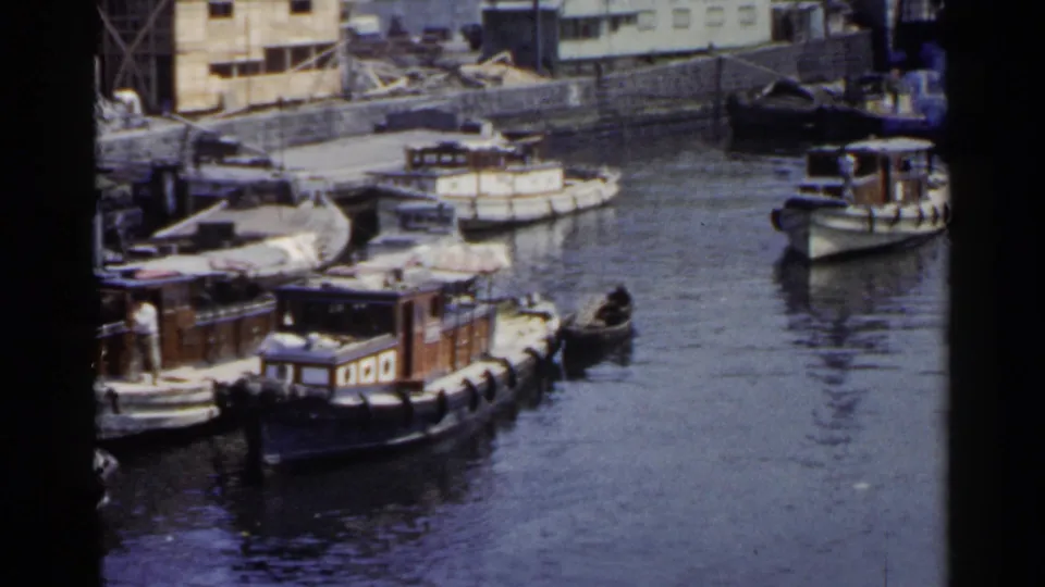 In this 1953 Japan port frame, a distant figure in light clothing appears near wooden boats tied beside docks and low buildings.