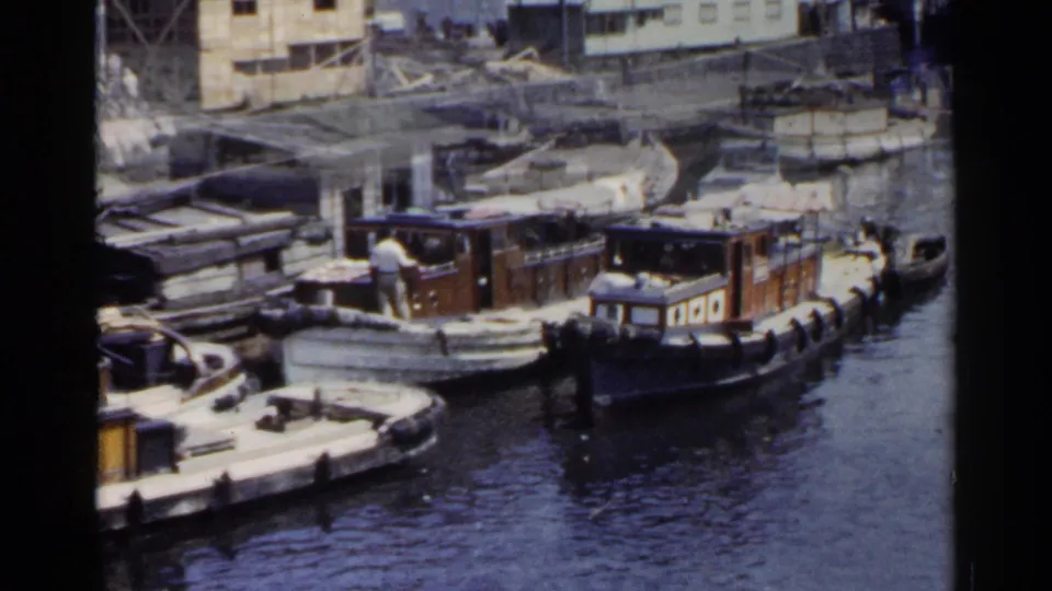 A 1953 Japan 8mm frame shows docked wooden boats, a distant person on one vessel, rippled water, and buildings behind the harbor edge.