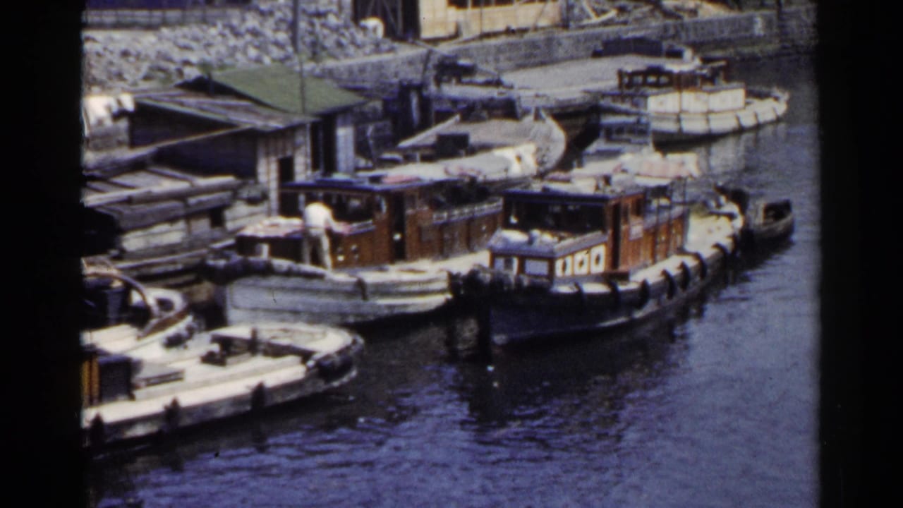 1953 Japan 8mm frame showing wooden boats tied along a stone quay, with men on deck and harbor water in the foreground.