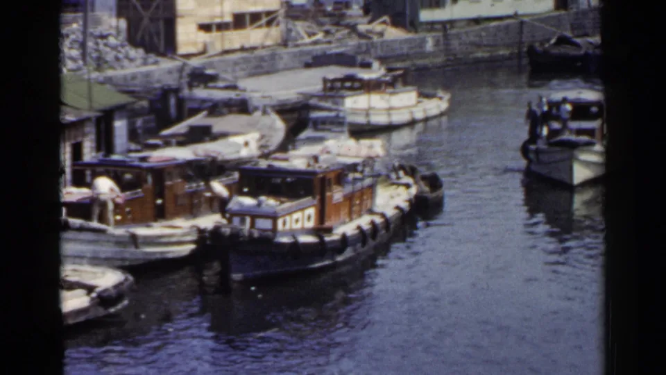 1953 Japan waterfront frame with moored wooden boats, men aboard, and another vessel appearing on the right side of the harbor.