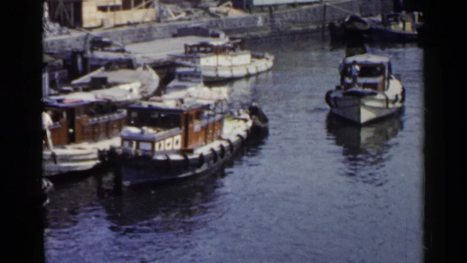1953 Japan 8mm view of docked wooden boats, workers near the decks, and a larger boat positioned to the right on calm harbor water.