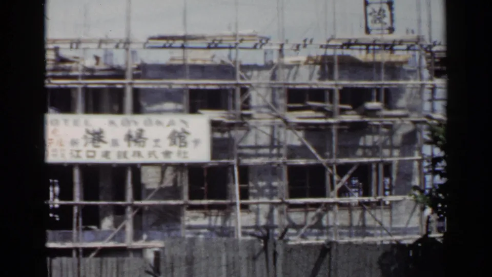 1953 Japan frame with slight motion blur, showing the scaffolded Hotel Koyokan construction frontage and surrounding metal framework.