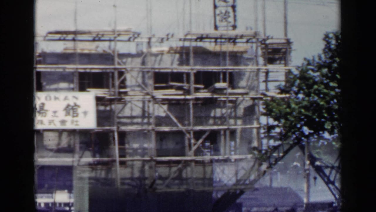 1953 Japan view of a building under construction, with scaffolding, Japanese lettering, and foliage at the frame edge.