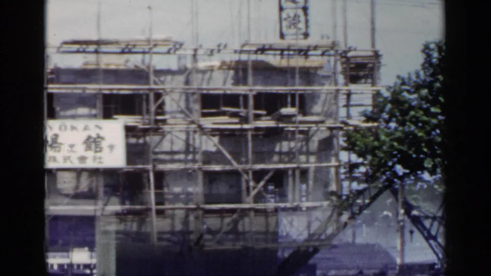 1953 Japan view of a building under construction, with scaffolding, Japanese lettering, and foliage at the frame edge.