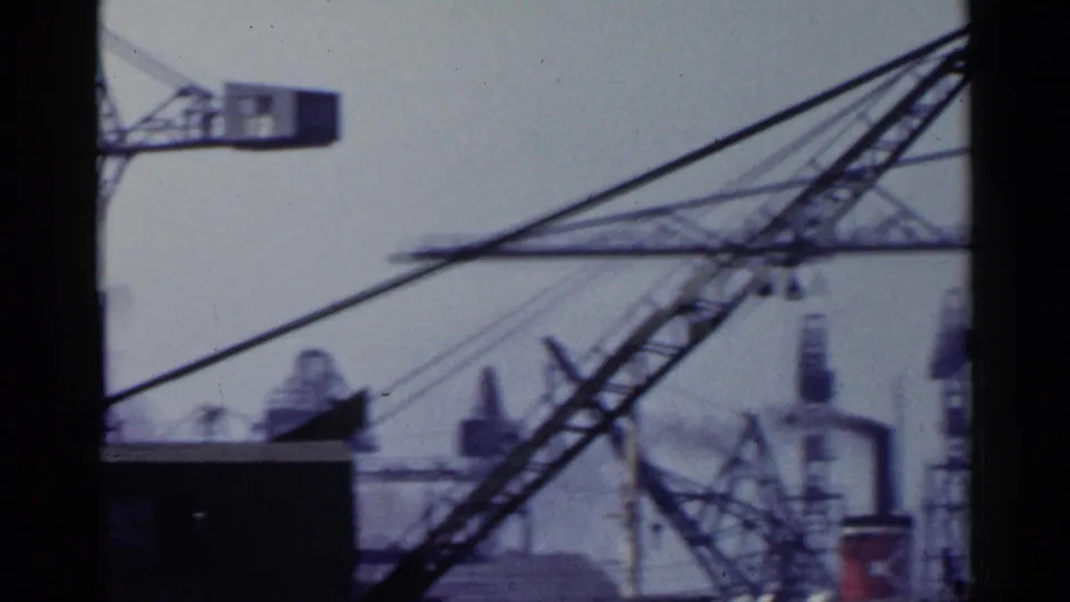 1953 Japan 8mm frame showing a diagonal crane boom and distant industrial port structures under a hazy sky.