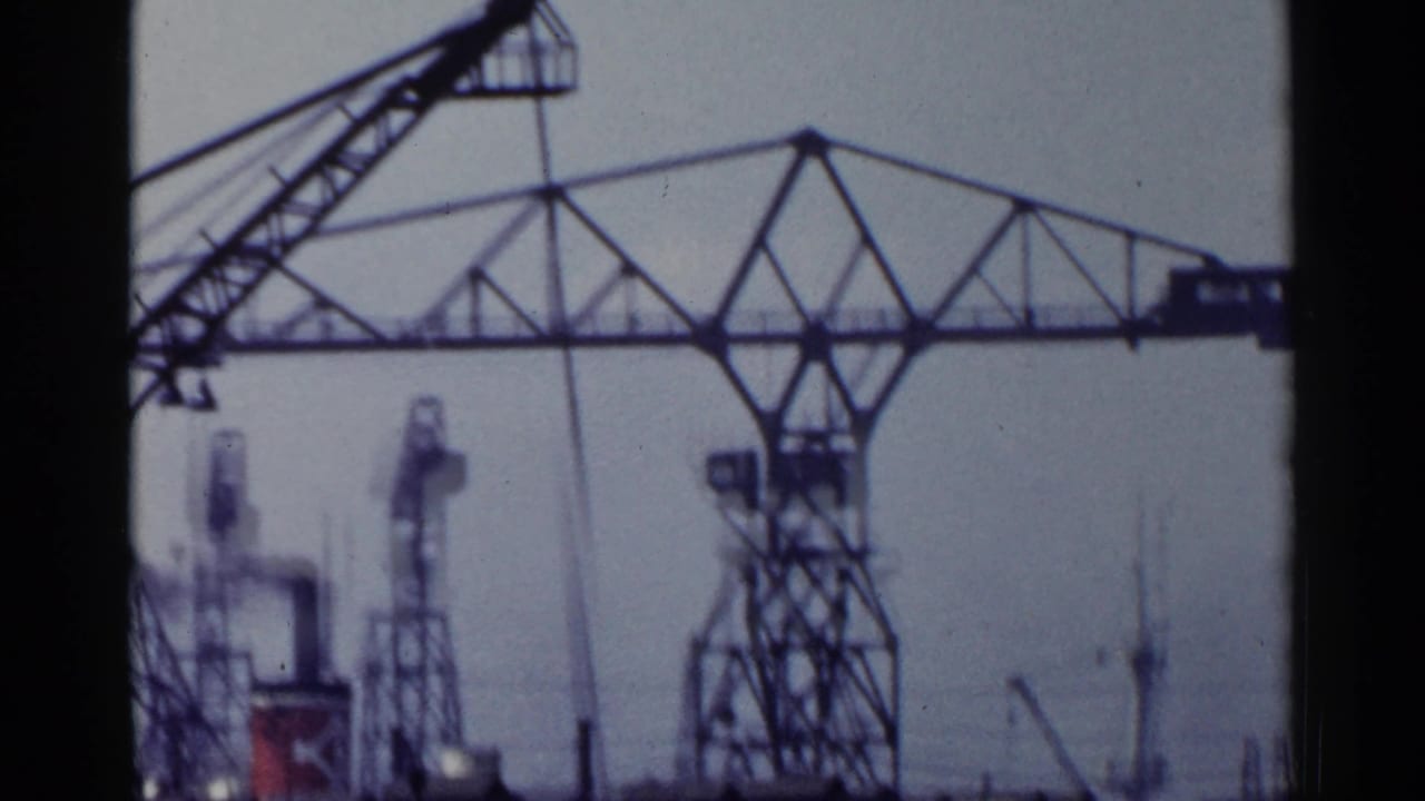1953 Japan 8mm frame showing tall harbor cranes and industrial framework near water, seen through soft grain and bluish film color.