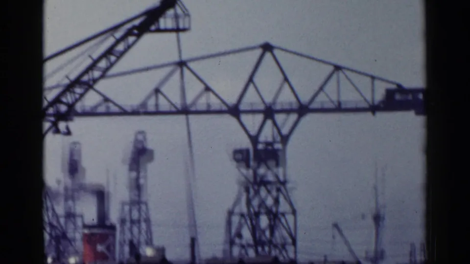 1953 Japan 8mm frame showing tall harbor cranes and industrial framework near water, seen through soft grain and bluish film color.
