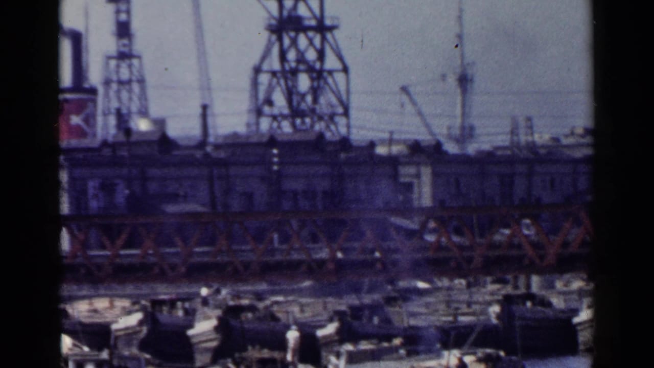 1953 Japan waterfront view with a red truss bridge, cranes, industrial buildings, and many small boats gathered below.