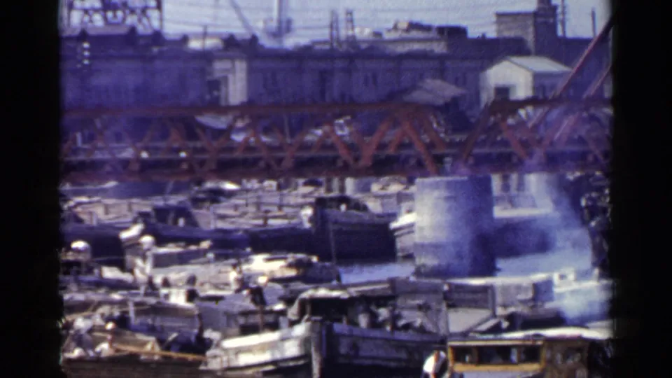 1953 Japan waterfront frame with dock workers, moored boats, a red truss bridge, and cranes or industrial structures in the background.