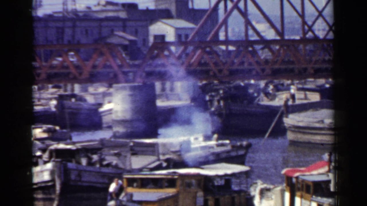1953 Japan 8mm frame showing a harbor beneath a red truss bridge, with small boats, smoke, and dock activity.