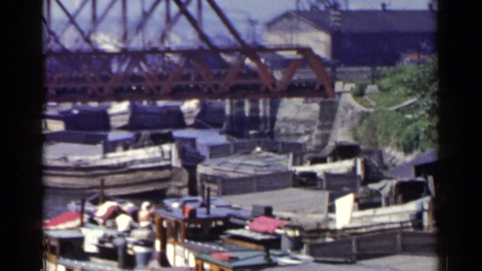 1953 Japan 8mm view of docked harbor craft, shoreline structures, and bridgework in a busy waterfront setting.