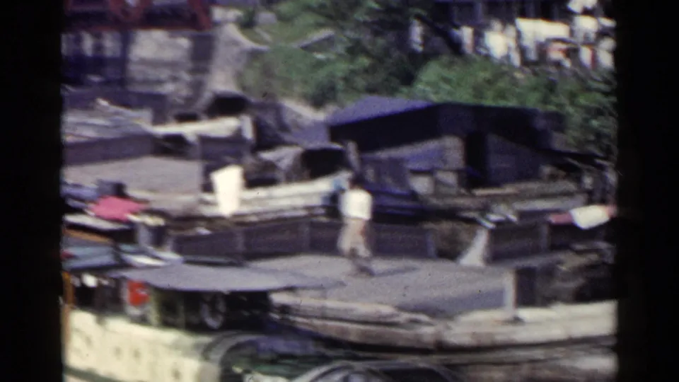 1953 Japan harbor frame with a man walking across a dock near moored boats, covered craft, and waterfront greenery.