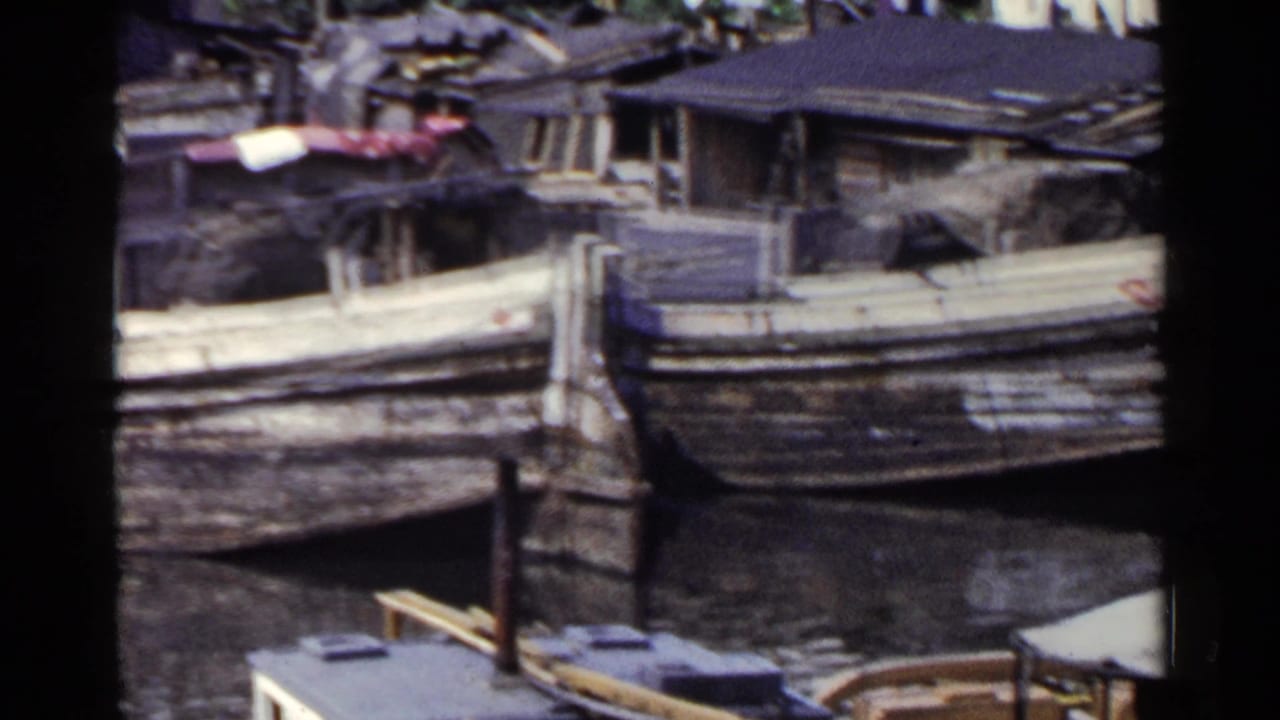 Wooden boats tied along a narrow waterfront with houses and posts nearby.