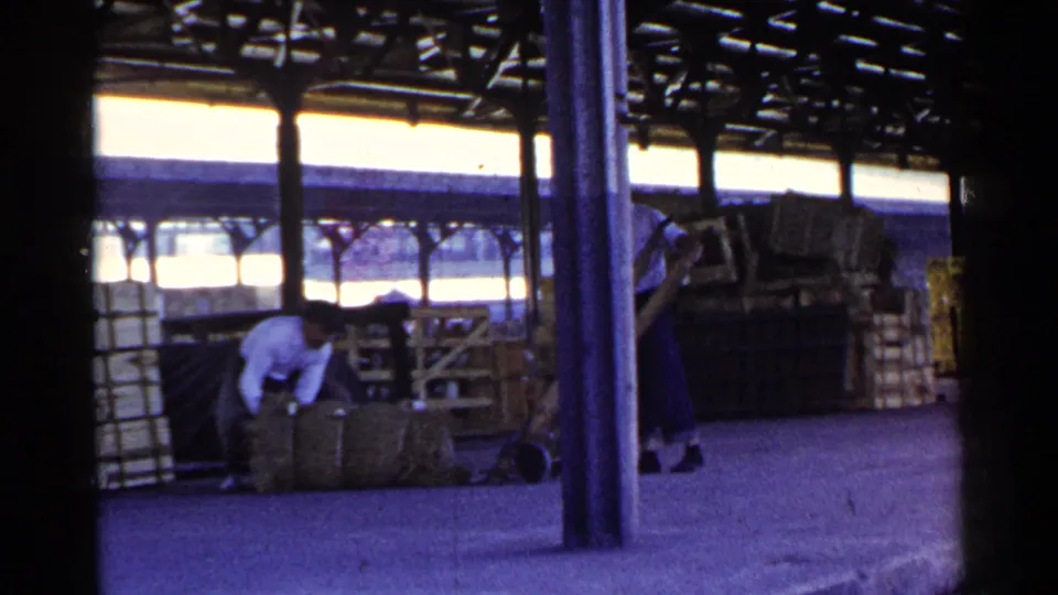 1953 Japan harbor work area with people handling cargo bundles under a roofed dock structure near boats.