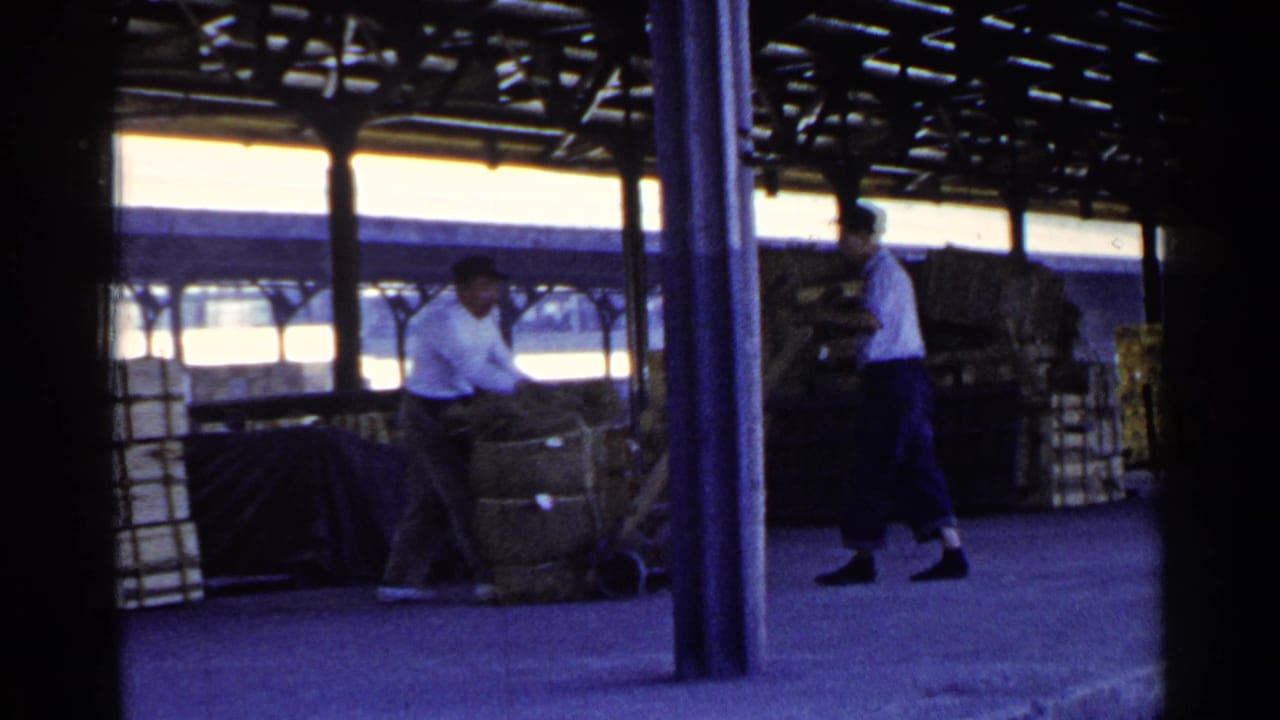 Two workers move a large bundle onto a hand cart on a covered dock.