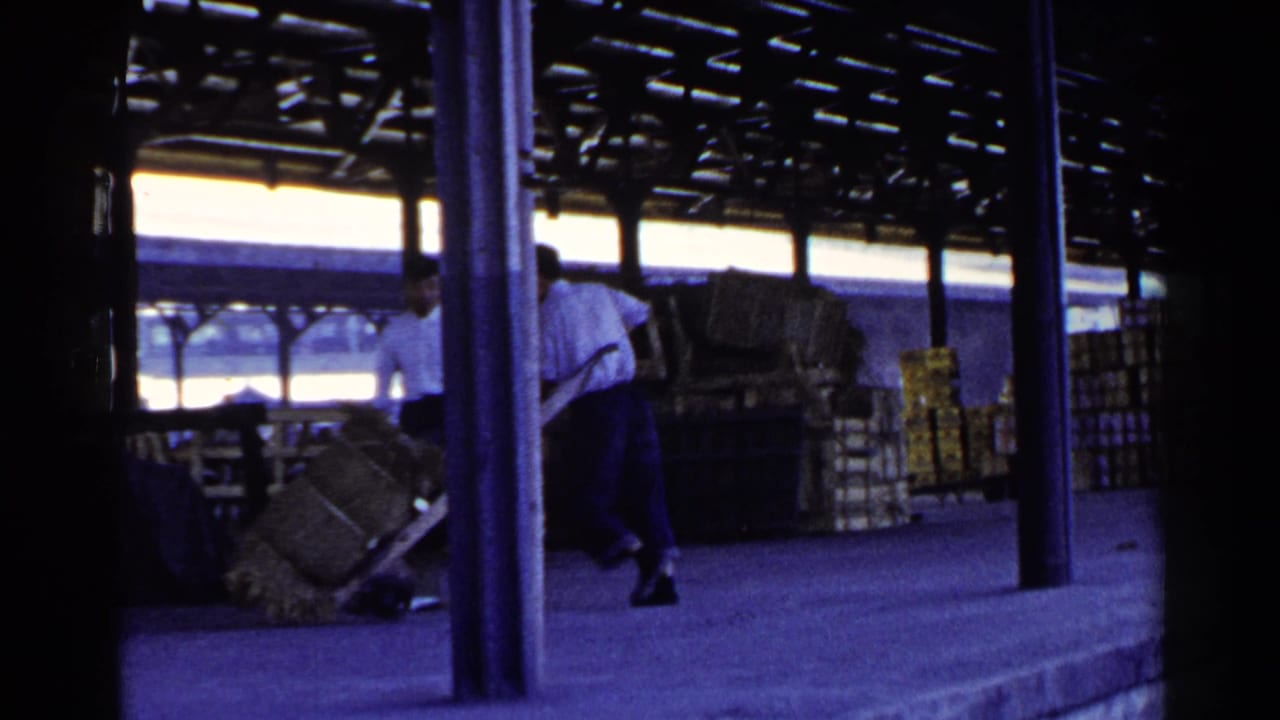 The dock workers continue shifting the bundle on the hand cart, with crates and roof supports visible behind them.