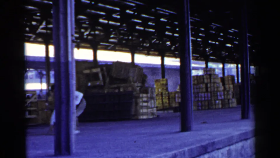 A worker bends near the edge of the frame beside the cart, with stacked cargo and columns across the covered dock.