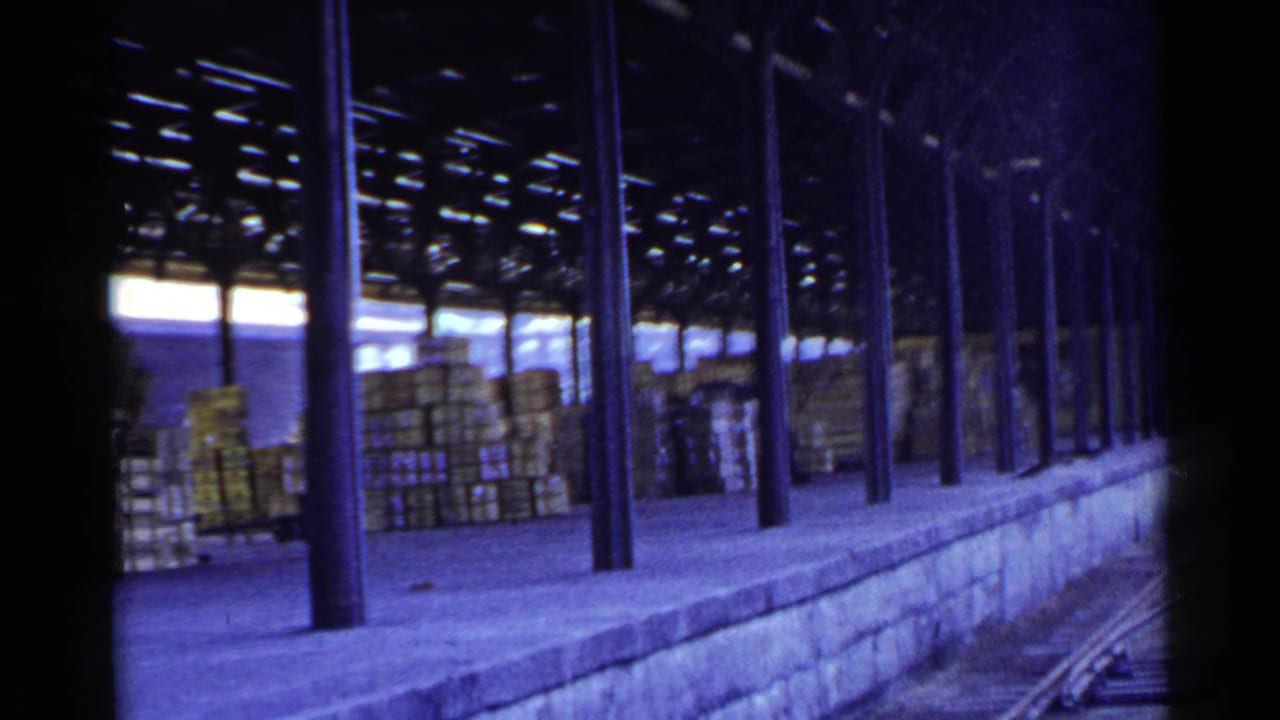 Passing 8mm view in Japan of a covered rail loading dock with stacked boxes and tracks along the platform.