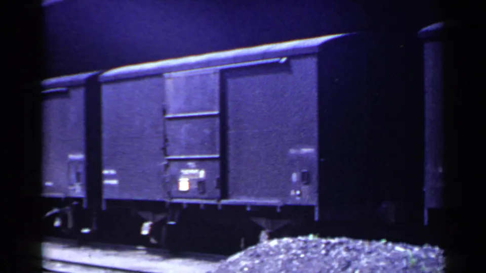 1953 Japan 8mm frame with a close view of dark freight cars, door panels, undercarriage wheels, and gravel beside the rails.