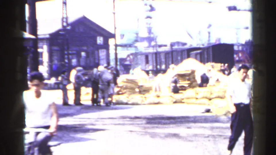 Daytime 1953 Japan rail-yard scene with workers and pedestrians near large bundled sacks beside a wooden station or yard building.