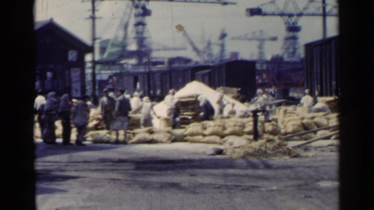 1953 Japan dockside view of men around cargo sacks, with train cars, cranes, and port structures visible behind them.