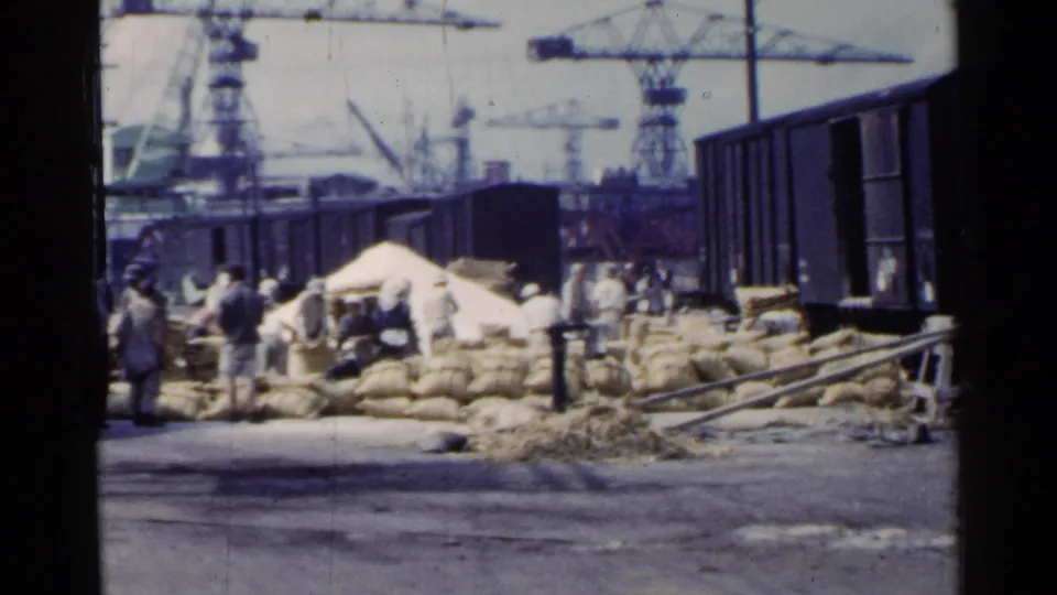 1953 Japan dock scene with stacked sacks, workers, a freight car, dark rail siding, and cranes across the port.