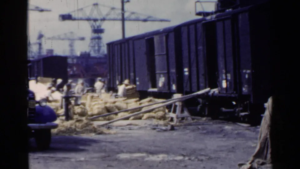 1953 Japan home-movie frame showing a blue truck edge, workers, sacks, dark freight cars, cranes, and rough ground.