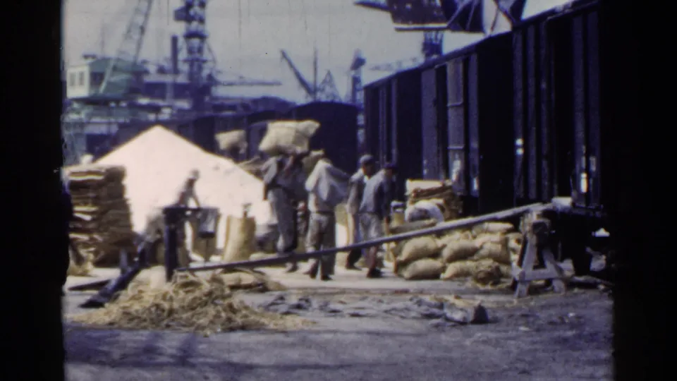 1953 Japan frame showing men near stacked sacks, rail cars, loading boards, cranes, and port-side freight activity.