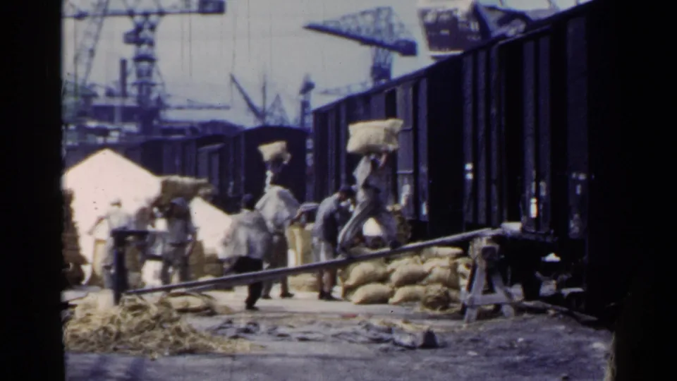 1953 Japan footage shows workers carrying burlap sacks near open freight cars, with cranes and piled cargo in the background.