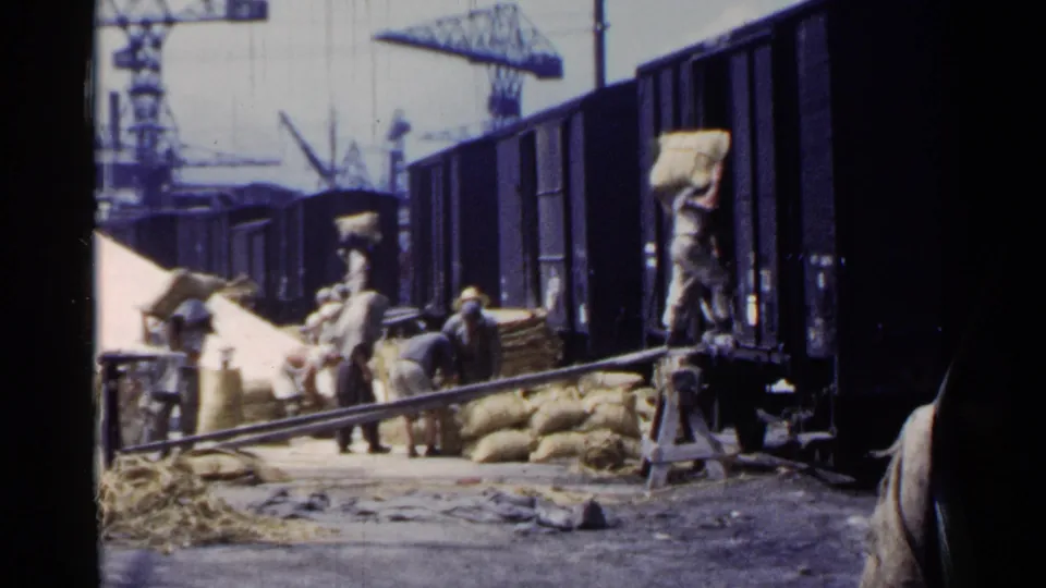 A mid-century dockside rail scene in Japan shows workers loading sacks by hand near open freight cars and stacked cargo.