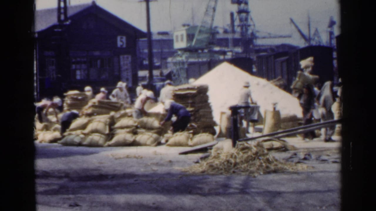 1953 Japan dockside scene with laborers moving sacks around a freight train, a pale cargo mound, cranes, and industrial sheds.