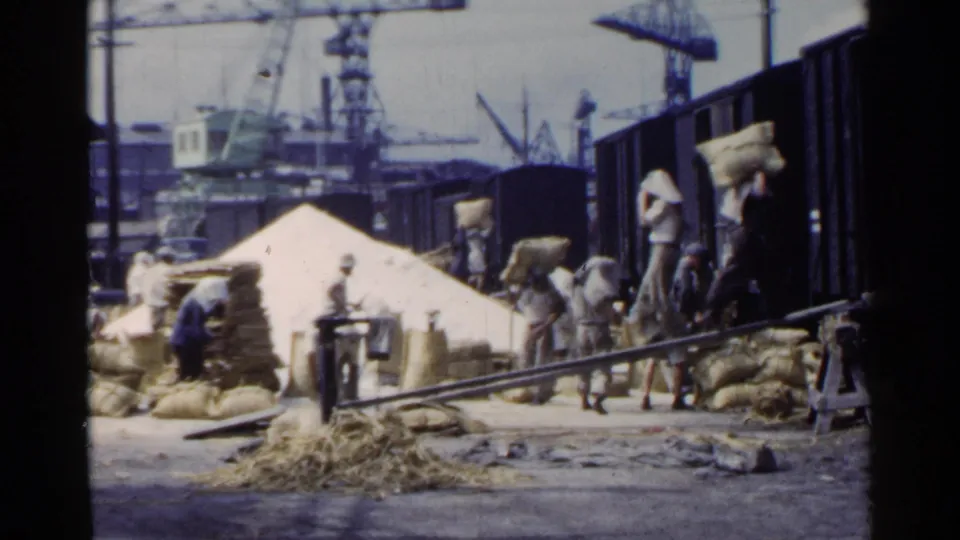 1953 Japan home-movie frame of workers loading sacks along freight railcars, with dock cranes and stacked cargo in the background.