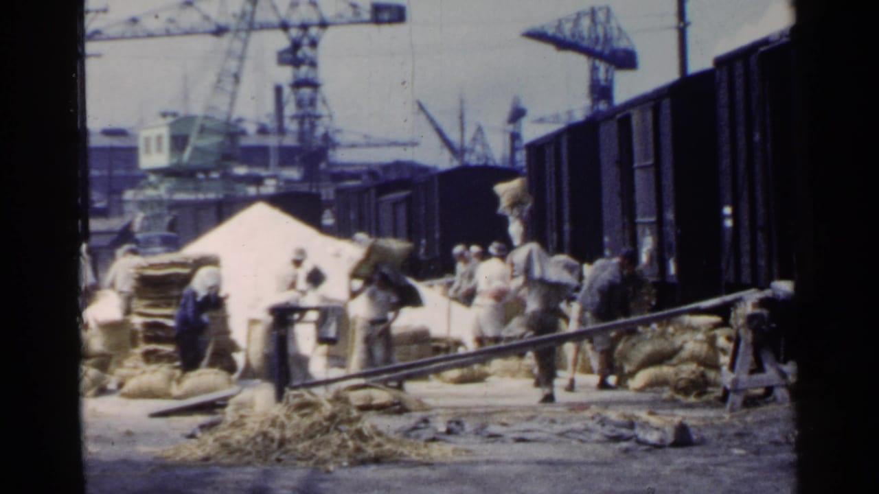 Workers carry sacks beside dark freight cars at an industrial dock with cranes in the background.