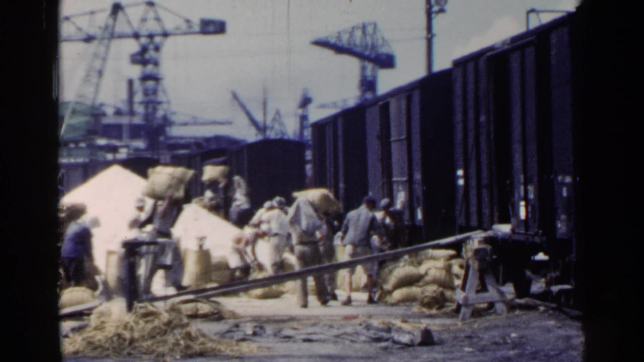 Workers in 1953 Japan appear to load or move sacks beside rail freight cars, with dock cranes and industrial equipment behind them.