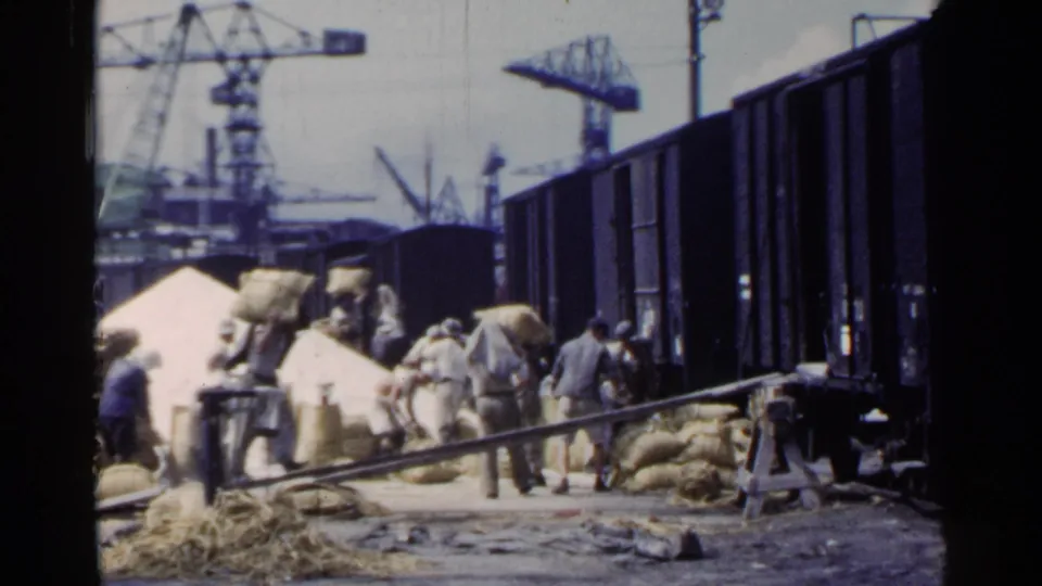Workers in 1953 Japan appear to load or move sacks beside rail freight cars, with dock cranes and industrial equipment behind them.