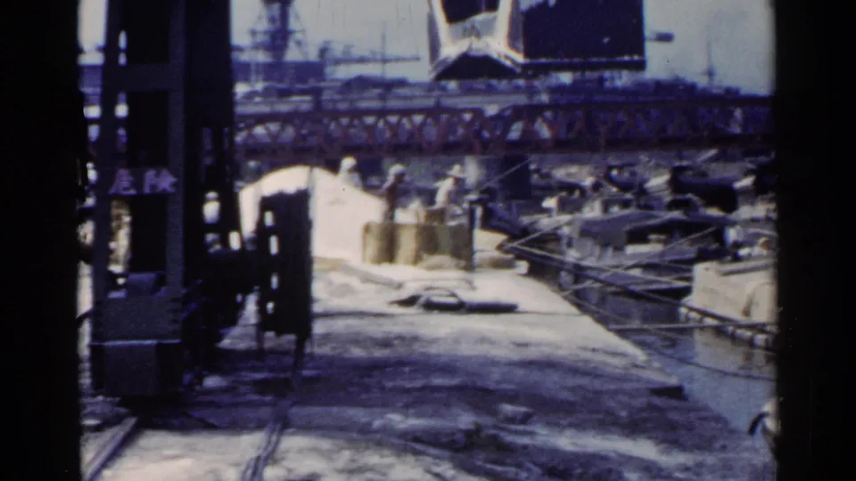 A crane bucket sweeps above a Japanese industrial waterfront, with boats, water, rail structures, and cranes visible in the 8mm frame.