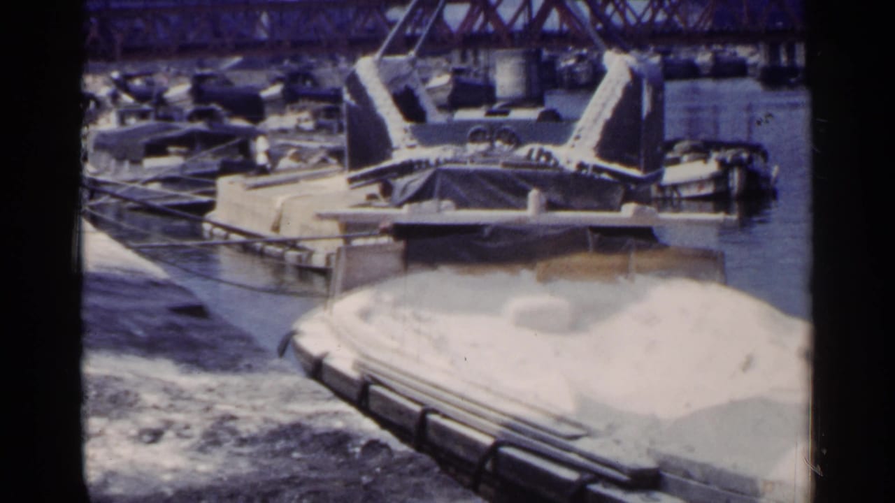 1953 Japan port view with a crane positioned over a barge carrying a white granular load, backed by small craft and a steel bridge.