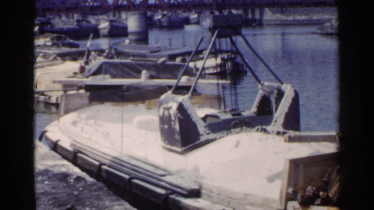 1953 Japan industrial port frame with a crane above a loaded barge, water and boats behind, and small distant people near the edge.