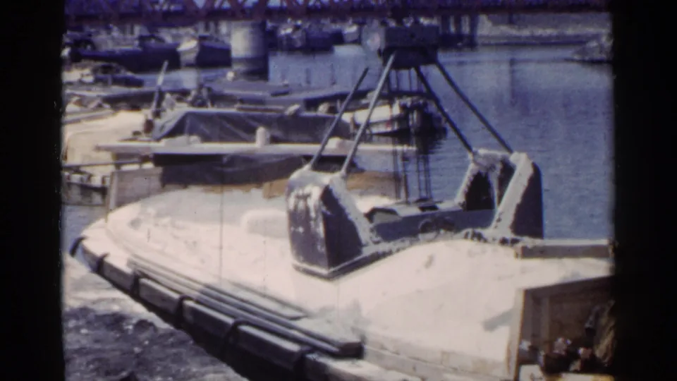 1953 Japan industrial port frame with a crane above a loaded barge, water and boats behind, and small distant people near the edge.