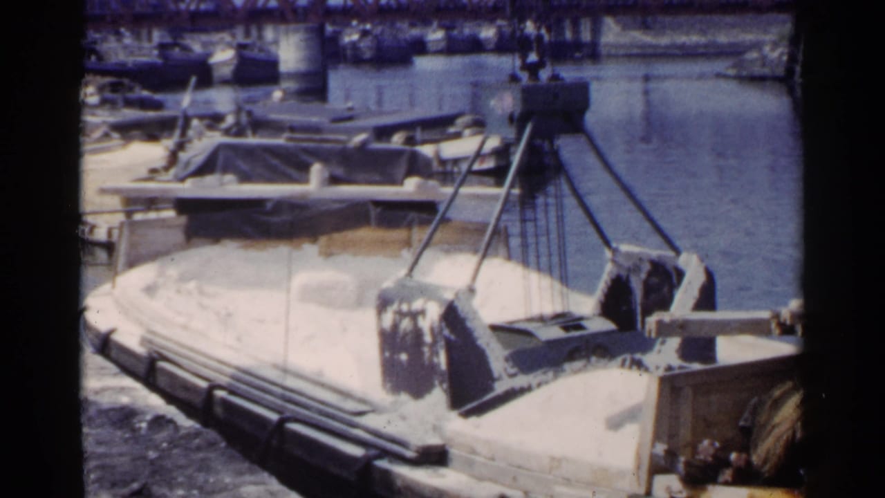 Crane rigging over a loaded barge on a harbor waterway with boats and a bridge behind.