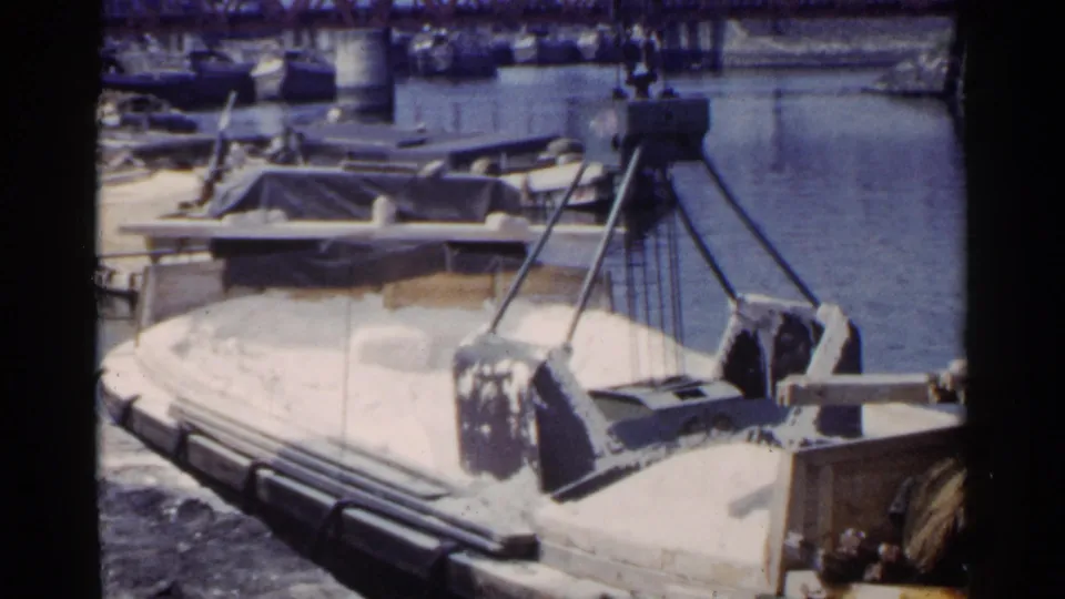 1953 Japan harbor view showing crane cables above a sand or gravel barge, with small boats and a truss bridge in the background.