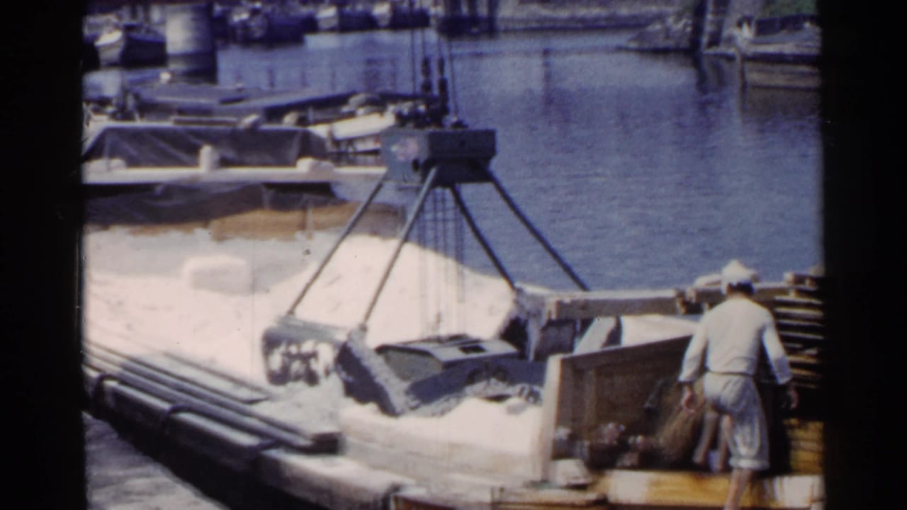1953 Japan port scene with a worker in white clothing beside stacked dock materials and a crane positioned over a loaded barge.