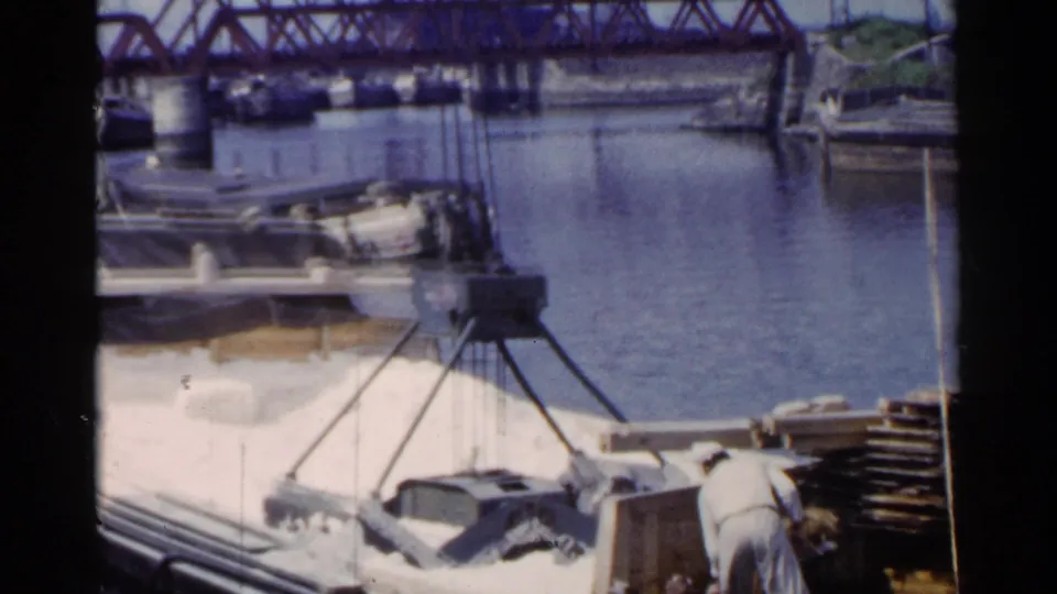 1953 Japan harbor footage showing a worker at the dock edge, crane rigging over a barge, and calm water beyond.