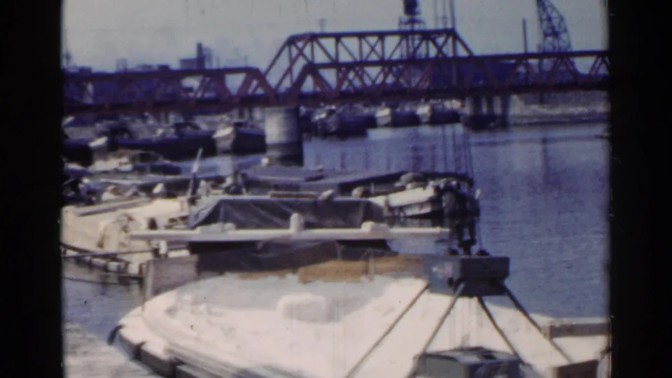 1953 Japan port view of a loaded barge beneath crane rigging, with moored boats, open water and a large truss bridge behind it.