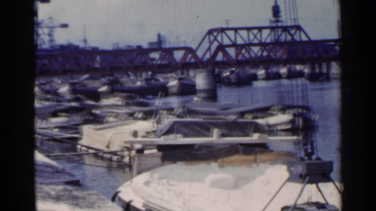 Boats moored at a dock with a red truss bridge and industrial cranes in the background.