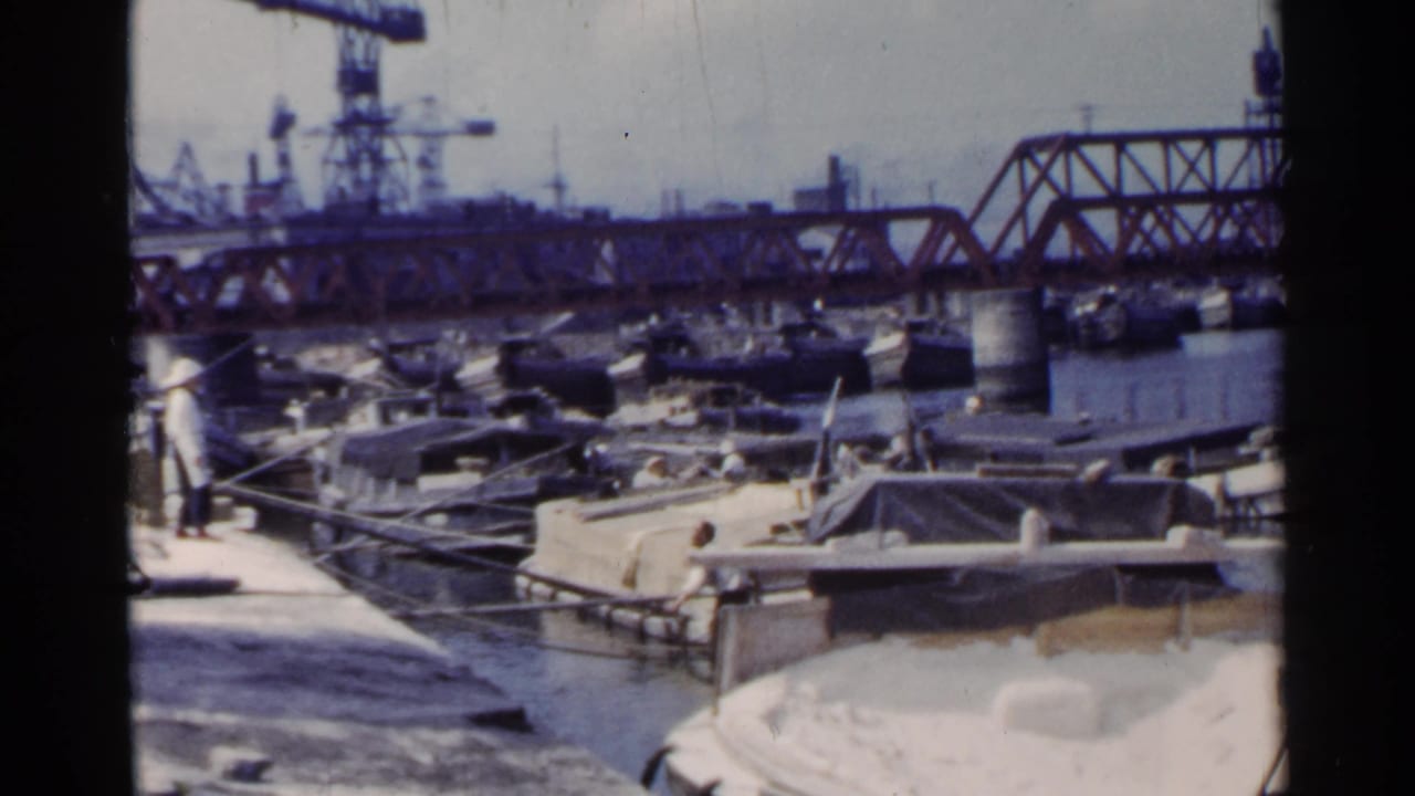 1953 Japan dock scene with workers near small boats, a red truss bridge over the water, cranes, and factory structures in the distance.